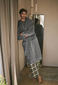 Homme en peignoir gris et pantalon de pyjama à carreaux, adossé à un rideau dans une chambre avec un grand miroir et des lampes suspendues.