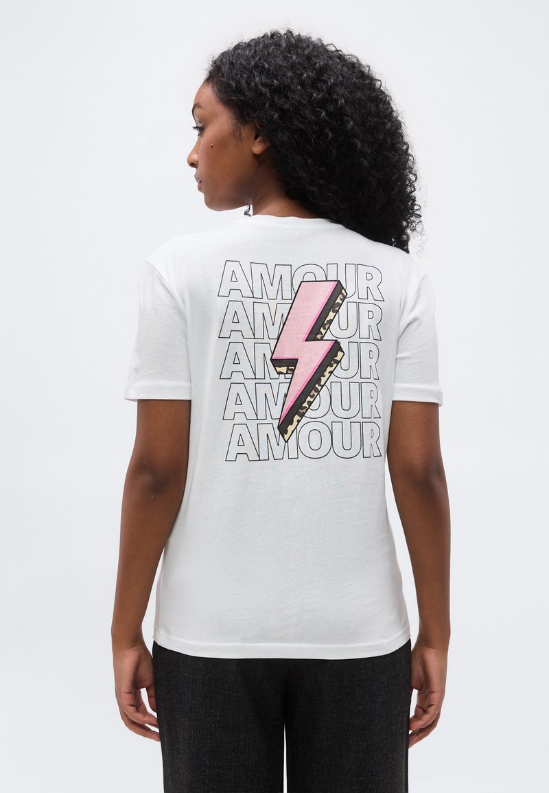 Γυναίκα με σγουρά μαλλιά που φοράει λευκό T-shirt με επαναλαμβανόμενο κείμενο «AMOUR» και μεγάλο ροζ σχέδιο κεραυνού στην πλάτη.