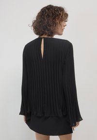 Persona con capelli ricci che indossa una blusa nera a maniche lunghe plissettata e una gonna nera, vista di spalle su uno sfondo neutro.