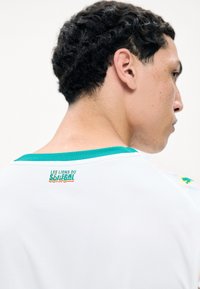 Jeune homme aux cheveux courts et bouclés portant un maillot de sport blanc avec un col turquoise et le texte « Les Lions du Sénégal » dans le dos.