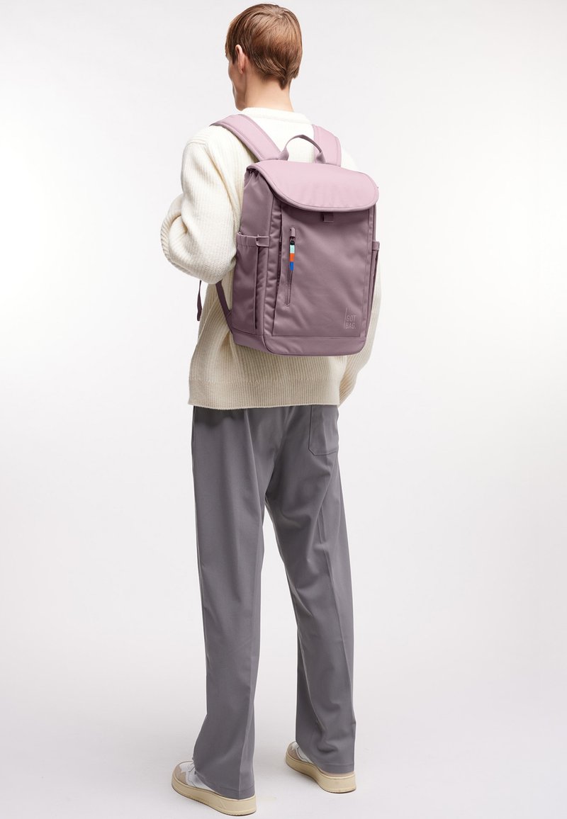 Personne portant un sac à dos violet clair avec des poches latérales, une fermeture éclair à l'avant, un pull beige, un pantalon gris et des baskets blanches, debout devant un fond blanc.
