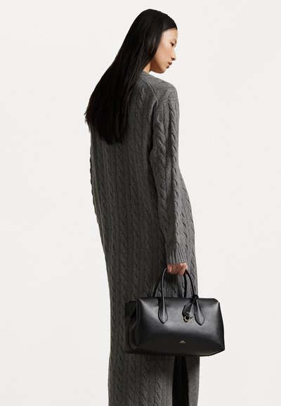 Μαύρη δερμάτινη τσάντα με κυλινδρικές λαβές, μινιμαλιστικός σχεδιασμός και χρυσή λεπτομέρεια λογότυπου. Το μοντέλο φοράει έναν μακρύ, γκρίζο πουλόβερ με πλέξη καλωδίου.