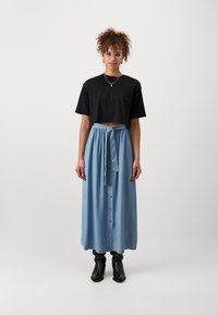 Camiseta corta negra combinada con una falda larga de mezclilla azul clara que presenta una tapeta de botones al frente y una cintura con lazo, complementada con botas negras hasta el tobillo.
