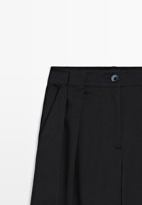 Pantalon noir avec une ceinture structurée, des plis et un bouton bleu. Le tissu a une texture lisse avec des poches latérales.