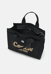 Mustat kankaiset kantokassit, joissa on kultaisella brodeerattu "Coach New York" -logo, kaksinkertaiset kahvat, vetoketjullinen tasku ja kangasvuori.
