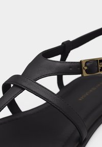 Μαύρο δερμάτινο σανδάλι με λεπτές σταυρωτές λουράκια και χρυσή αγκράφα στο λουράκι του αστραγάλου.