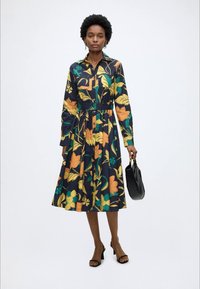 Femme portant une robe longue à manches longues, longueur genou, avec de grands motifs floraux jaunes, orange et verts, tenant un sac à main noir et portant des talons noirs.