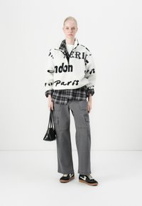 Hvid fleece-pullover med sort tekst, lagd over en sort og grå ternet skjorte. Grå cargo-bukser og sorte sneakers med gummisåler.