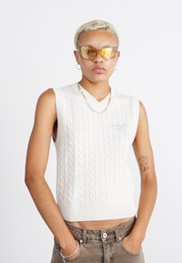Creme farge kabelstrikket uten ermer genser med V-hals, med lys blå brodert tekst. Brukt med lyse jeans og solbriller.