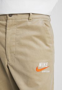 Khaki kordfljolbyxor med en broderad orange Nike-logotyp och vit text, med en slät yta och klassisk passform.