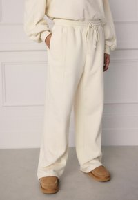 Cream-colored, relaxed-fit sweatpants with an elastic waistband and drawstring. Pockets on the sides, paired with tan loafers.
