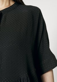 Czarna bluzka o fakturze z delikatnym, wypukłym wzorem, z krótkimi rękawami i okrągłym dekoltem, wykonana z lekkiego materiału.