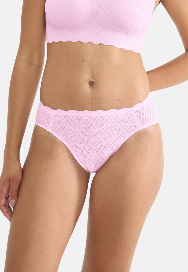 Γυναίκα που φοράει ροζ ανοιχτό ροζ δαντελένιο σουτιέν και ταιριαστό εσώρουχο με υψηλή γραμμή και κυματιστά άκρα, μπροστά σε απλό φόντο.