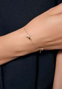 Złota bransoletka łańcuszkowa z małymi urokami w kształcie serca i motyla, połączona delikatnym ogniwem, noszona na dłoni na tle teksturowanego czarnego tła.