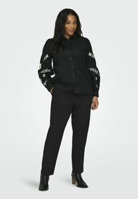 Melns krekliņš ar apdrukātiem ziedu dizainiem uz piedurknēm, apvienots ar melnām biksēm un augstpapēžu potīšu zābakiem. Zīdainas auduma tekstūra.