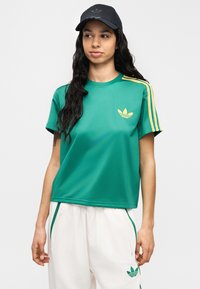 Πράσινο αθλητικό μπλουζάκι με κίτρινες ρίγες, στρογγυλή λαιμόκοψη και μικρό λογότυπο στο στήθος. Συνδυασμένο με λευκό παντελόνι με πράσινες λεπτομέρειες.