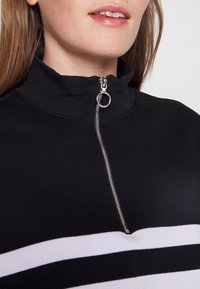 Top negro con cremallera y cuello alto, que presenta un hardware de cremallera plateado y rayas blancas horizontales en el pecho. Textura de tela suave.