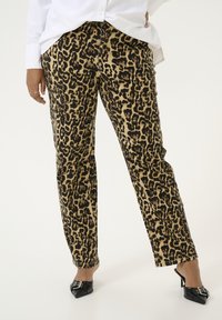 Luipaardprint broeken met een tan en zwart patroon, relaxed fit en opgerolde boorden, gecombineerd met zwarte puntige schoenen en een witte blouse.