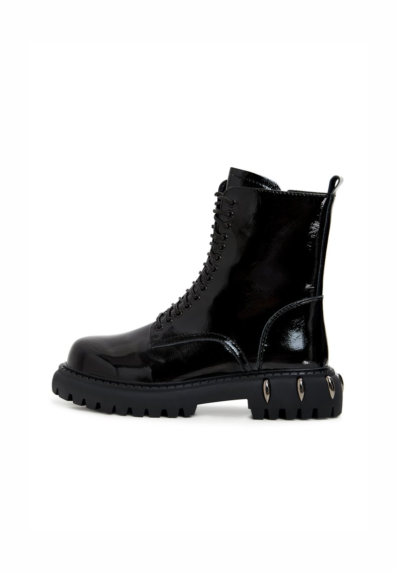 Musta lakattu nilkkabotti paksulla mustalla pohjalla, pyöreällä varvalla ja metallikoristeilla pohjassa. Varustettu nauhakiinnityksellä.