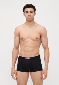 Fekete boxer alsónadrág széles gumis derékrésszel, amelyen a "BOSS" márkaneve fehér színnel szerepel. Sima anyagú és testhezálló kialakítású.