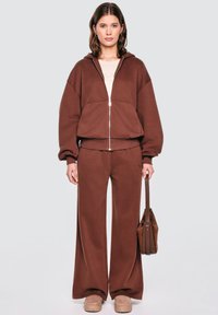 Brązowy zip-up hoodie z kapturem, połączony z szerokimi brązowymi spodniami dresowymi. Model trzyma brązową torbę; miękka faktura, casualowy design.