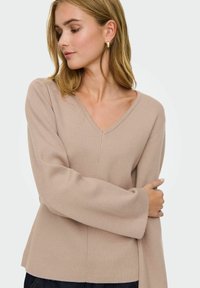 Femme aux cheveux blonds mi-longs portant un pull beige à col en V et des boucles d'oreilles en or, regardant vers le bas avec les bras croisés.