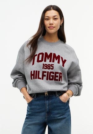Frau mit langen dunklen Haaren, die einen grauen Pullover mit rotem Tommy Hilfiger 1985 Schriftzug, blaue Jeans und eine goldene Uhr trägt, Hände in den Taschen.