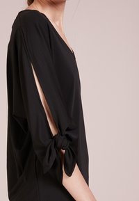 Svart topp med V-ringning, med trekvartsärmar med knytaccenter och sidöppningar, tillverkad av ett mjukt, lätt material.