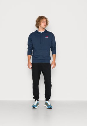 Sudadera navy con bolsillo delantero y capucha ajustable, combinada con pantalones de chándal negros y zapatillas multicolores. Fondo plain.