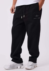 Persona che indossa pantaloni sportivi neri con un piccolo logo sulla coscia destra e sneakers bianche, in piedi contro uno sfondo semplice.