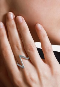 Silberner Zickzackring, getragen an einer Hand mit natürlichen Nägeln. Glatte, glänzende Oberfläche. Hautfarbe ist hell, mit einer glatten Textur.