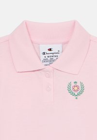 Lys rosa polo skjorte med en myk tekstur, med krage, tre knapper og et blomstermønster med en krone i grønt og rosa.