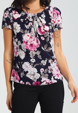 Vrouw draagt een marineblauw, kortemouws bloesje met roze, witte en grijze bloemenpatronen, gecombineerd met een zwarte broek, toont armtatoeages.