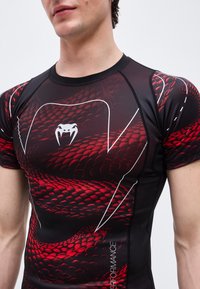 Κοντινό πλάνο ενός ατόμου που φοράει μαύρο και κόκκινο συμπιεστικό μπλουζάκι με μοτίβο λέπι φίδι και λευκό λογότυπο με δόντια φιδιού στο στήθος.