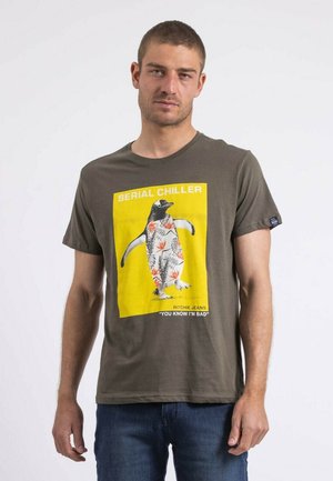 T-shirt vert olive avec un imprimé graphique jaune d'un pingouin dans un feuillage tropical, avec le texte "SERIAL CHILLER" et "YOU KNOW I'M BAD."