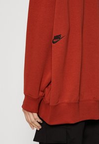 Mikina v rezavě červené barvě s měkkou texturou, s černým logem Nike na rukávu a vroubkovanými náplety. Střih v ležérním stylu.