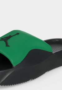 Κοντινό πλάνο ενός πράσινου και μαύρου σαγιοναριού με μαύρο λογότυπο Jumpman στο λουράκι και υφασμένο μαύρο πέλμα.