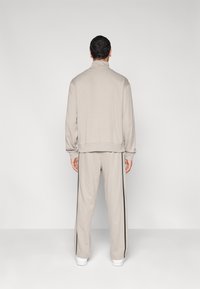 Φωτεινή μπεζ φόρμα με φούτερ που κλείνει με φερμουάρ και παντελόνι με ελαστική μέση. Διαθέτει μαύρες ρίγες στο πλάι και απαλό, υφασμάτινο υλικό με υφή.