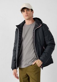 Giovane uomo con berretto beige, giacca a cappuccio nera sopra una maglietta grigia e pantaloni oliva, in piedi contro uno sfondo bianco semplice.