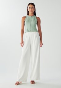 Top senza maniche verde menta con zip frontale, abbinato a pantaloni bianchi larghi con dettagli di tasche laterali. Tacchi aperti di colore neutro.