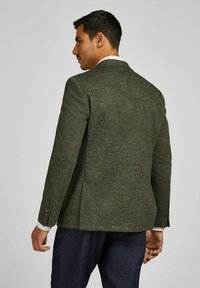 Man in een getextureerde donkergroene tweed blazer met knopen op de mouwen, in combinatie met donkerblauwe broek, staand met zijn rug naar de camera tegen een effen achtergrond.
