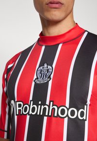 Punase ja musta triibulise jalgpallipluusiga, millel on kõrge krae, tikkitud klubi vapp ja suur valge nimi "Robinhood" esiküljel.