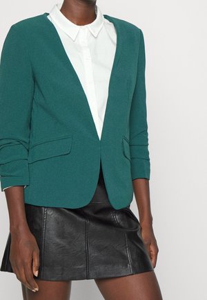 Vrouw draagt een donkergroen blazer over een wit bloesje met knopen en een zwarte leren minirok, staande tegen een effen lichte achtergrond.