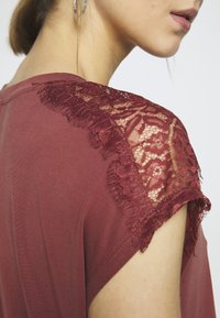 Bordová tričko s krátkými rukávy, které má složité krajkové detaily na rameni, jemnou texturu a volný střih.