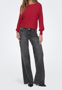 Pull tricot rouge à côtes avec un col large, associé à un jean noir taille haute, ample, et des escarpins noirs élégants.