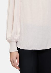 Blusa rosa chiaro con maniche voluminose, polsini a costine e un tessuto morbido, leggermente texturizzato. Abbinata a pantaloni neri aderenti.