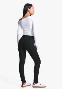 Fekete, skinny farmernadrág hátsó zsebekkel, fehér hosszú ujjú felsővel párosítva. A modellen fehér sportcipő van barna részletekkel.