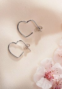 Orecchini a cerchio a forma di cuore in argento con una finitura liscia, dotati di chiusure a farfalla, su uno sfondo di morbido tessuto pastello.