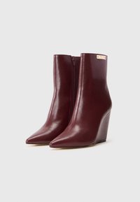 Bottines à talon compensé en cuir bordeaux avec un bout pointu et une fermeture éclair sur le côté. Présente une plaque logo métallique sur la tige. Texture lisse.