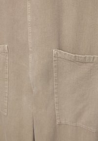 Beiger Stoff mit matter Textur, ausgestattet mit zwei vorderen Taschen und sichtbarer Naht. Einfaches, klares Design mit leicht zerknittertem Aussehen.